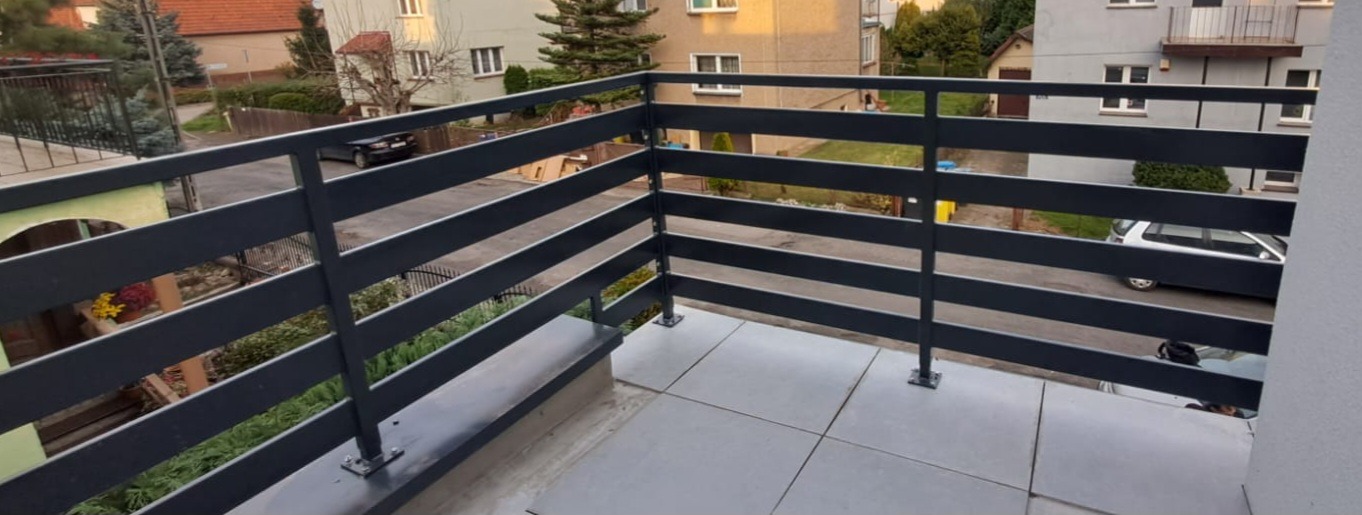 Nowoczesna, pozioma balustrada balkonowa w kolorze grafitowym, zamontowana na jasnych płytkach w Wysocku. Widok na ulicę i zabudowę mieszkalną.