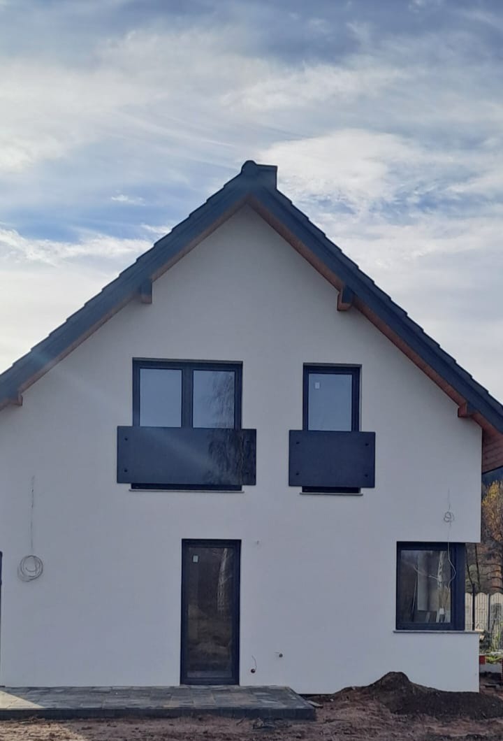 Nowoczesny dom jednorodzinny z czarnymi, minimalistycznymi balustradami balkonowymi i stolarką okienną w kolorze antracytu, na tle błękitnego nieba.