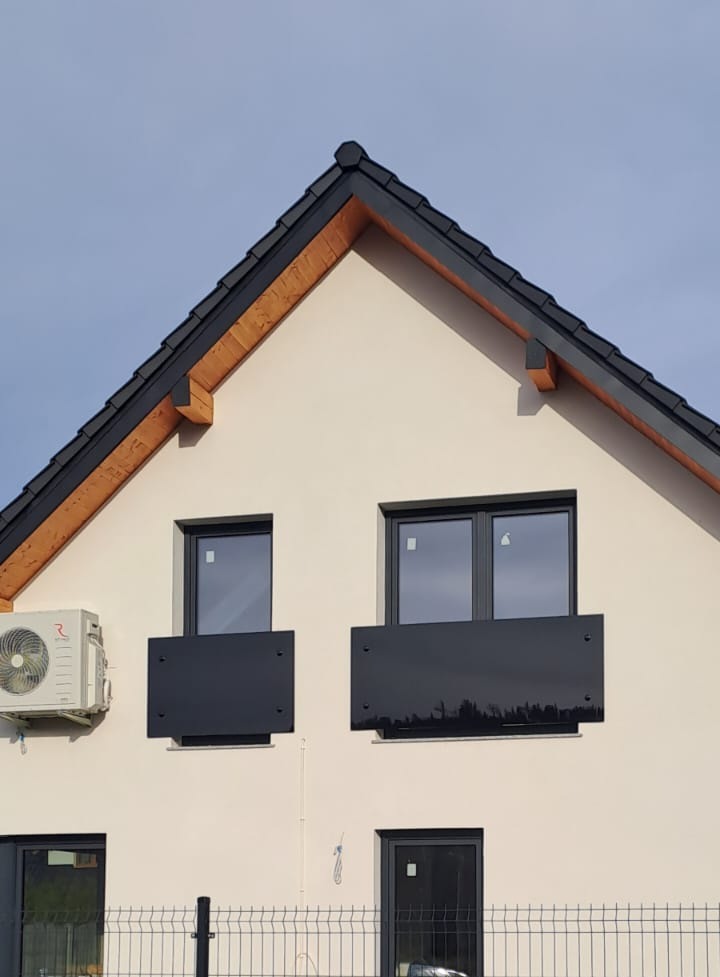 Elewacja domu z czarnymi, metalowymi balkonami bez barierki, widoczne okna i klimatyzator. Ujęcie z dołu, na pierwszym planie fragment ogrodzenia.