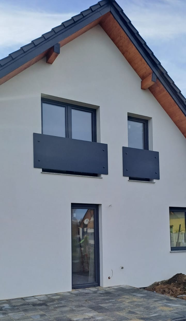 Elewacja domu z ciemnymi, prostokątnymi osłonami balkonowymi, zamontowanymi na dwóch oknach na piętrze. Widoczna kostka brukowa na ziemi i fragment dachu.