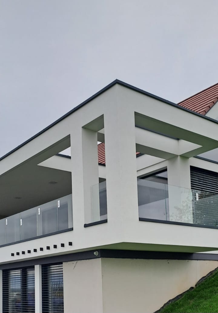 Nowoczesny balkon z przeszkloną balustradą i białymi, geometrycznymi elementami konstrukcyjnymi. Widok z dołu, fragment dachu z czerwoną dachówką w tle.