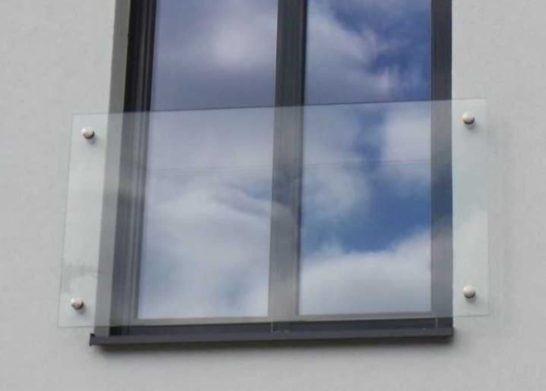 Szklana osłona okienna zamontowana na zewnątrz budynku, przymocowana za pomocą czterech metalowych śrub. W szybie odbija się błękitne niebo z chmurami.