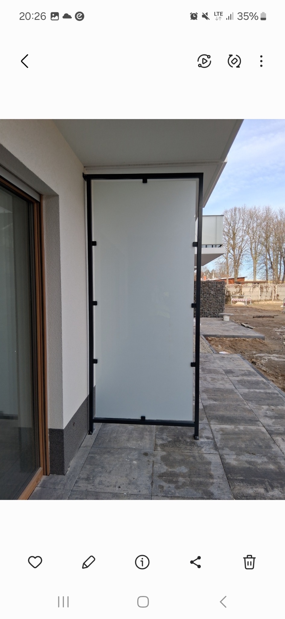 Szklana balustrada balkonowa z czarną ramą, widok z boku, na tle kostki brukowej i częściowo odsłoniętego terenu budowy. Minimalistyczny design.