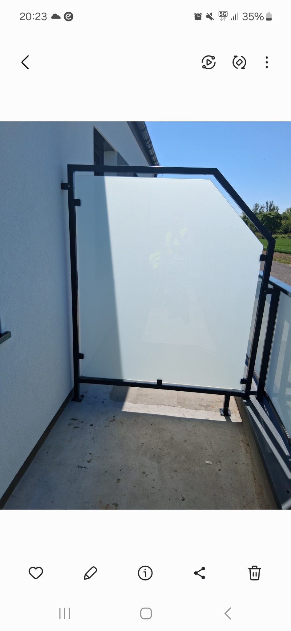 Nowoczesna, matowa balustrada szklana z czarną ramą na balkonie. Nietypowy kształt szkła, widok na zieleń w tle. Minimalistyczny design.