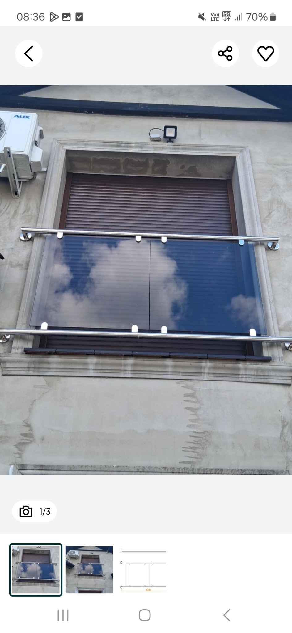 Szklana balustrada balkonowa z metalowymi poręczami, odbijająca błękitne niebo z chmurami. Widok z dołu na balkon z brązową roletą i klimatyzatorem na szarej elewacji.