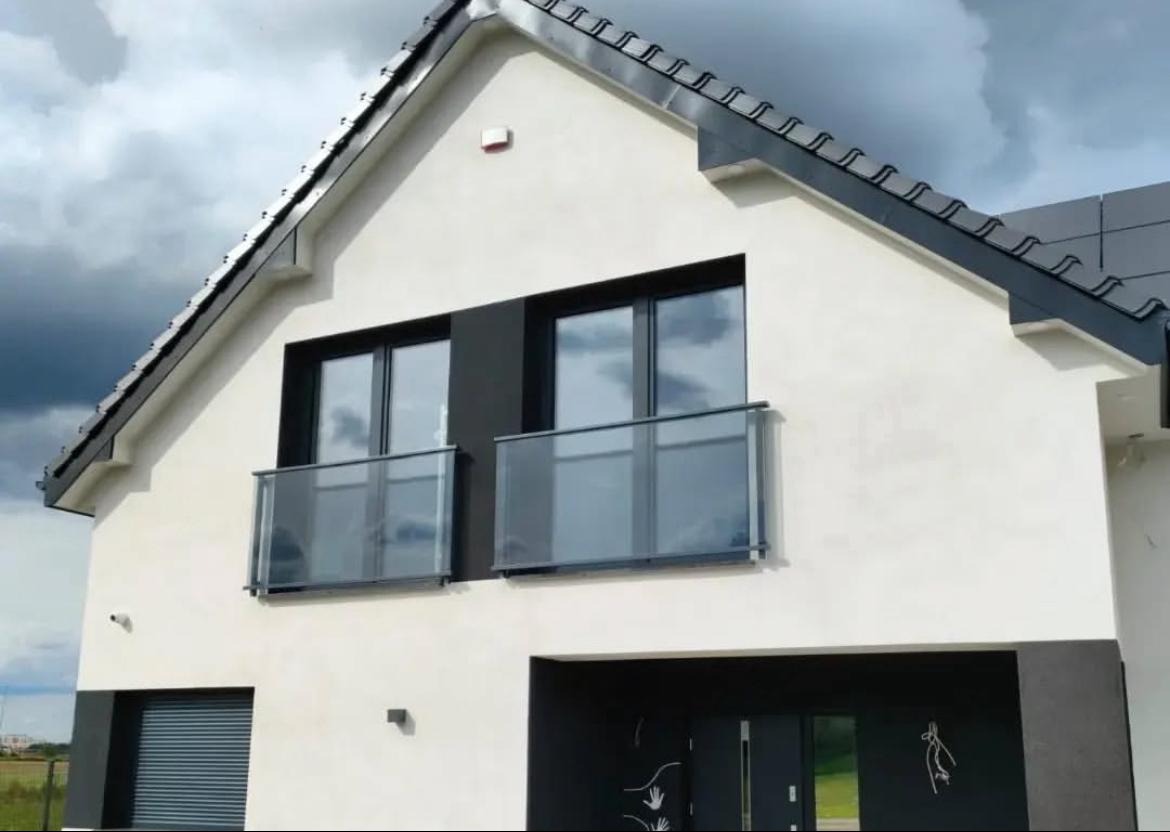 Nowoczesny dom z dwoma balkonami z balustradami szklanymi, czarne ramy okienne i drzwi, biała elewacja z szarymi akcentami, pochmurne niebo w tle.