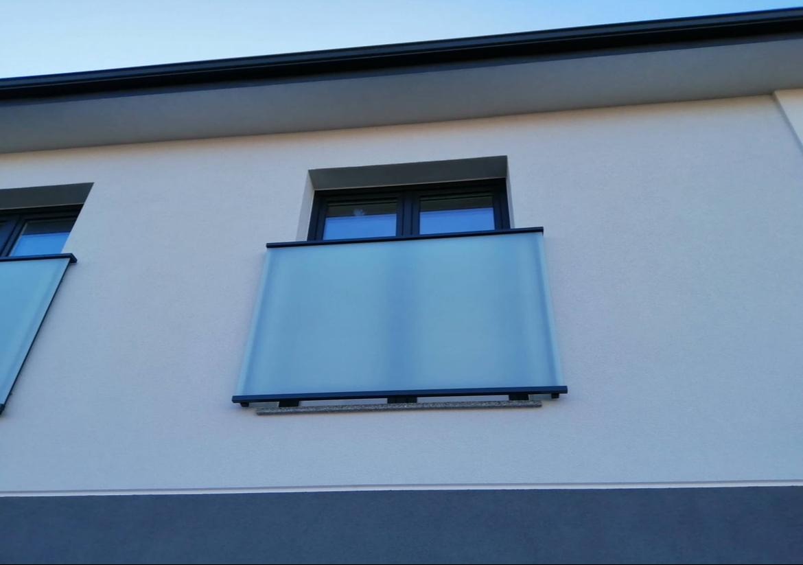 Nowoczesna balustrada szklana z czarnymi ramami, zamontowana na balkonie budynku o jasnej elewacji. Widok z dołu, podkreślający minimalistyczny design i przejrzystość szkła.