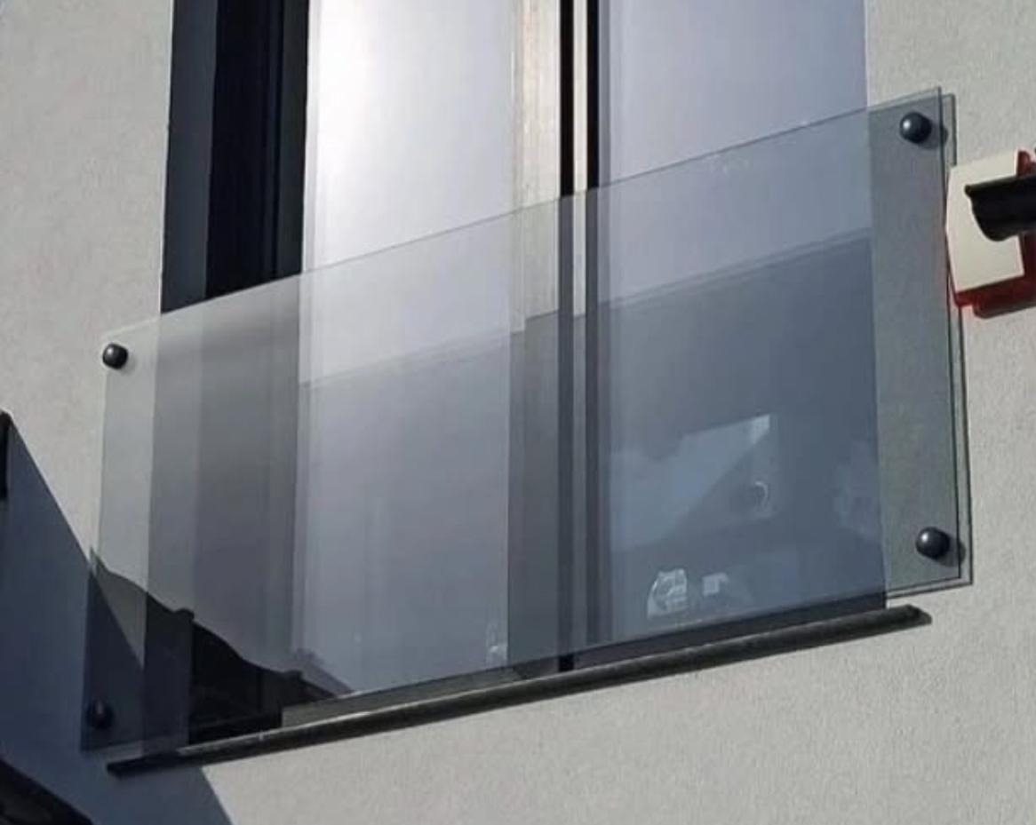 Nowoczesna, szklana balustrada balkonowa z ciemnymi mocowaniami, widoczna z bliska, z refleksami światła na szkle i fragmentem okna w tle. Minimalistyczny design.