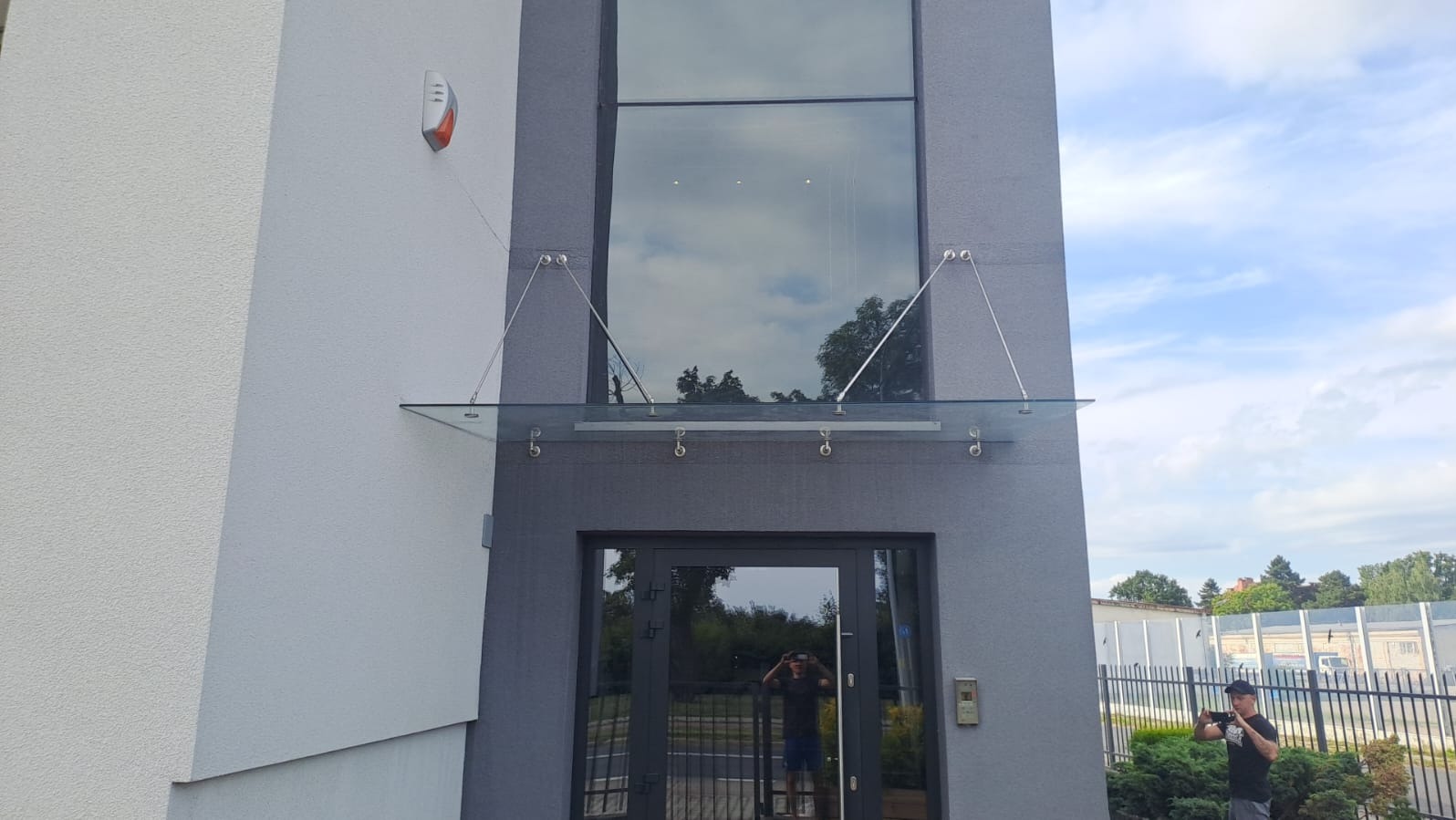 Szklany daszek nad wejściem do budynku, wsparty na metalowych linkach. Nowoczesny design, widoczne mocowania. W tle błękitne niebo i zieleń drzew.