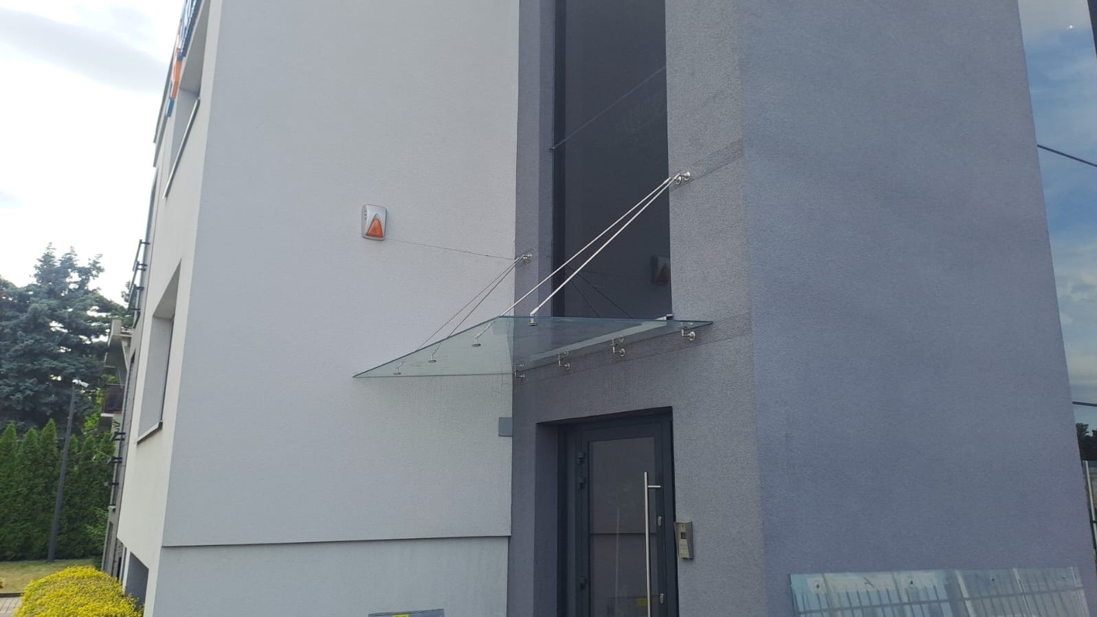 Szklany daszek nad wejściem do budynku, wsparty na stalowych linkach. Nowoczesny design, minimalistyczna konstrukcja, szara elewacja budynku.