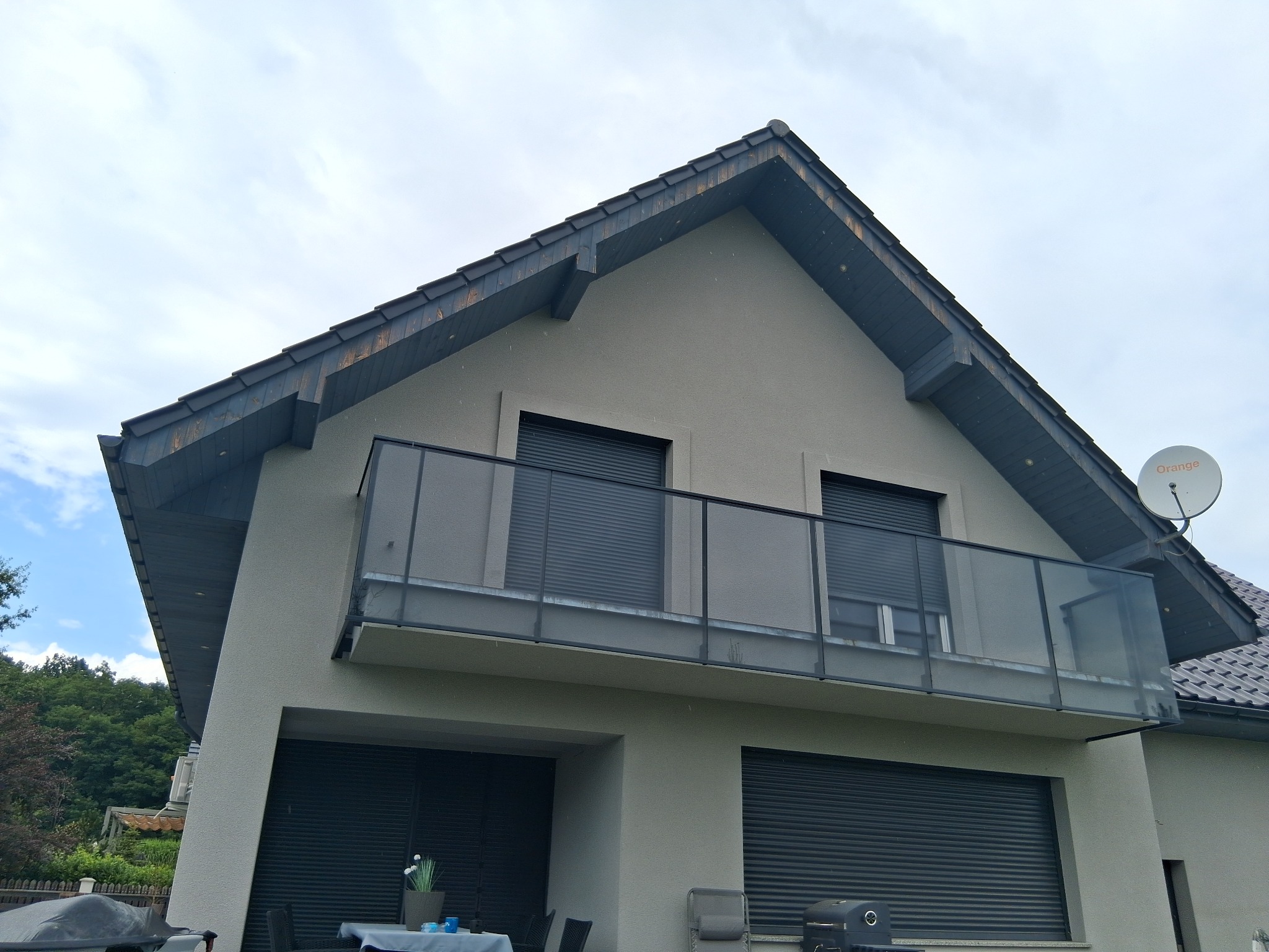 Nowoczesny dom z balkonem zabezpieczonym szklaną balustradą, widok z dołu na elewację i dach z ciemną dachówką. Na dachu antena satelitarna.