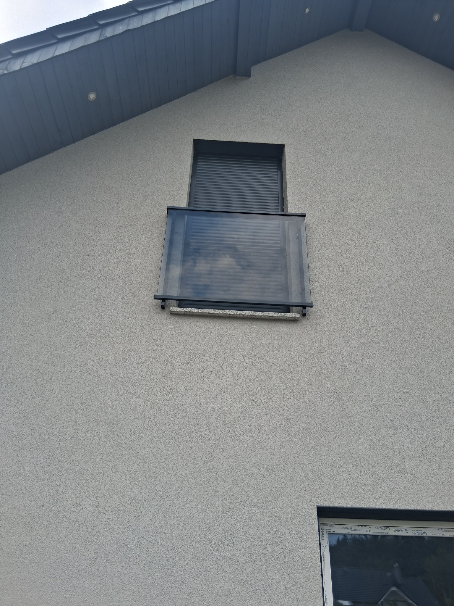 Szklana balustrada balkonowa z szarymi elementami mocującymi na tle jasnej elewacji budynku z oknem z roletą. Widok z dołu, neutralne oświetlenie.