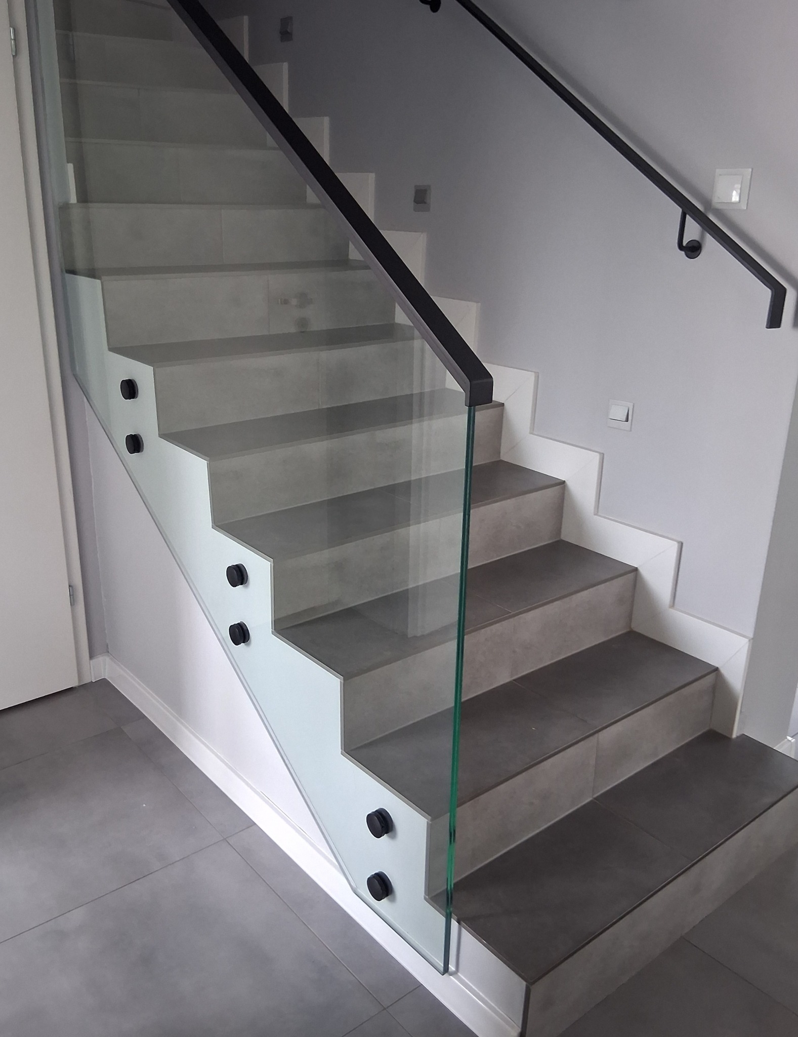 Nowoczesne schody z szarą okładziną, zabezpieczone szklaną balustradą z czarnymi mocowaniami i poręczą. Minimalistyczny design, jasne wnętrze.