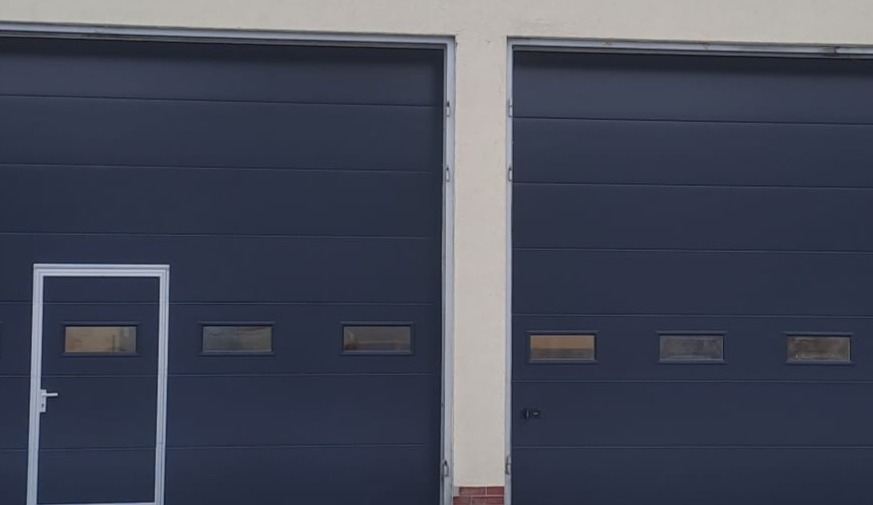 Dwie segmentowe bramy przemysłowe w odcieniach szarości z przeszkleniami i drzwiami serwisowymi po lewej stronie. Solidna konstrukcja, estetyczne wykończenie.