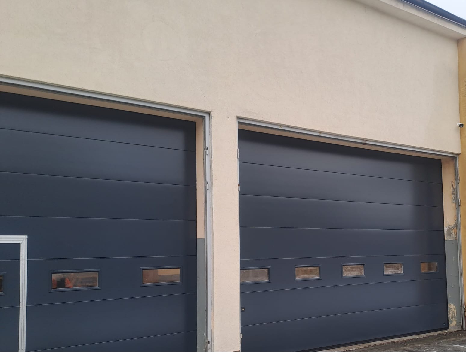 Dwie segmentowe bramy garażowe w kolorze antracytowym z poziomymi panelami i małymi okienkami, zamontowane w beżowej elewacji budynku.