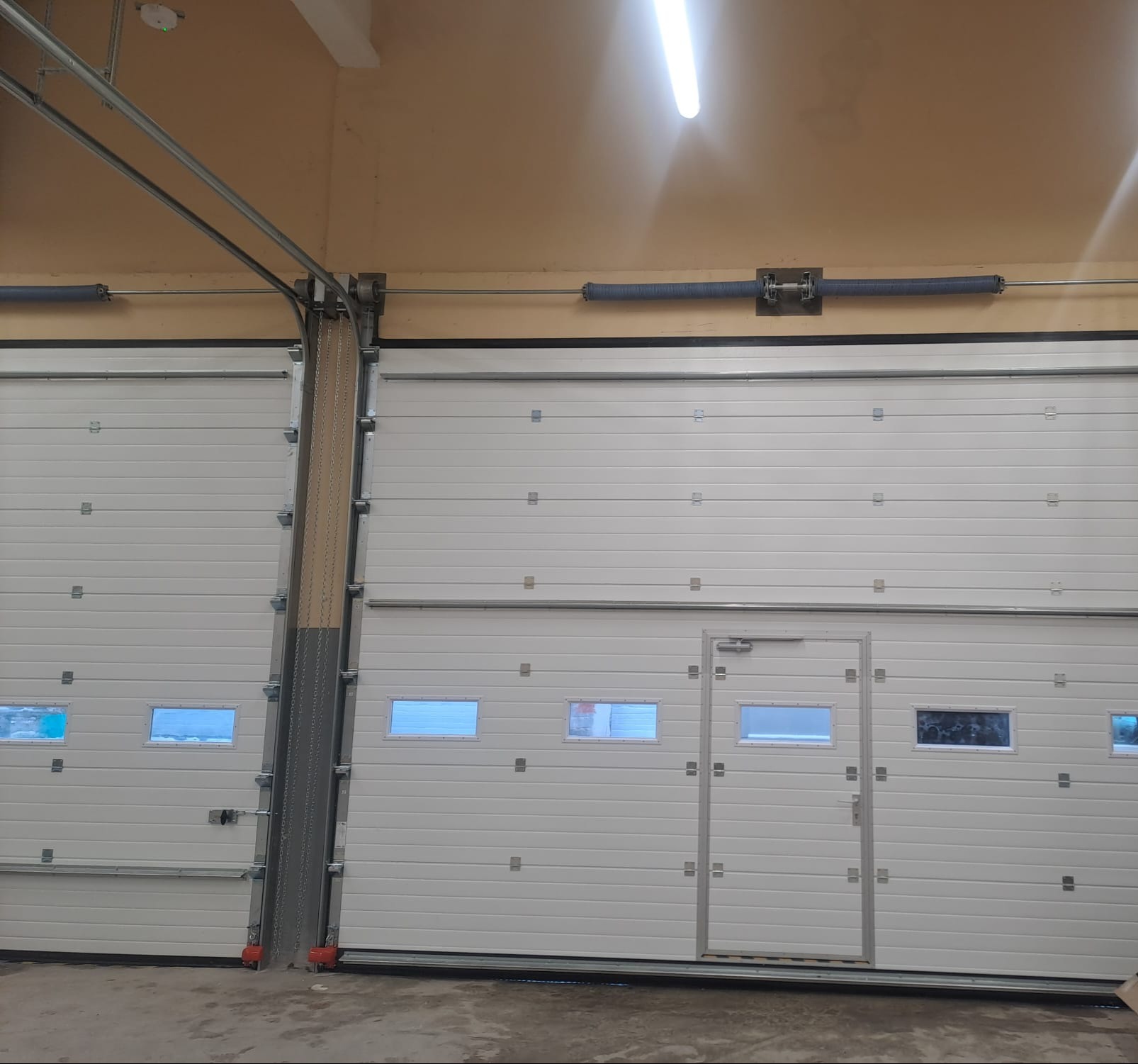 Dwie białe, segmentowe bramy garażowe z oknami, widoczne mechanizmy sprężynowe i prowadnice. Jedna brama z drzwiami przejściowymi. Oświetlenie sufitowe.
