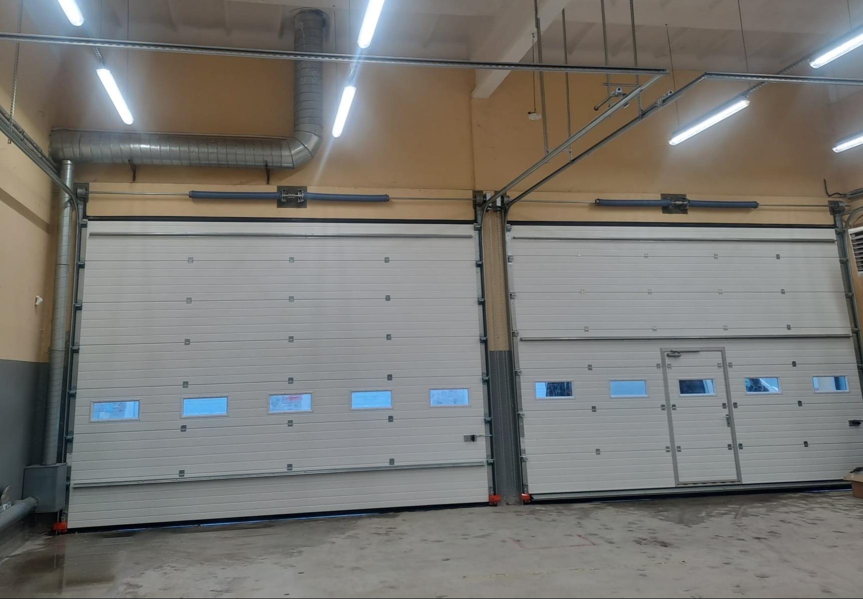 Dwie białe, segmentowe bramy garażowe z oknami, jedna z drzwiami wejściowymi, w hali z widoczną instalacją wentylacyjną pod sufitem i oświetleniem jarzeniowym.