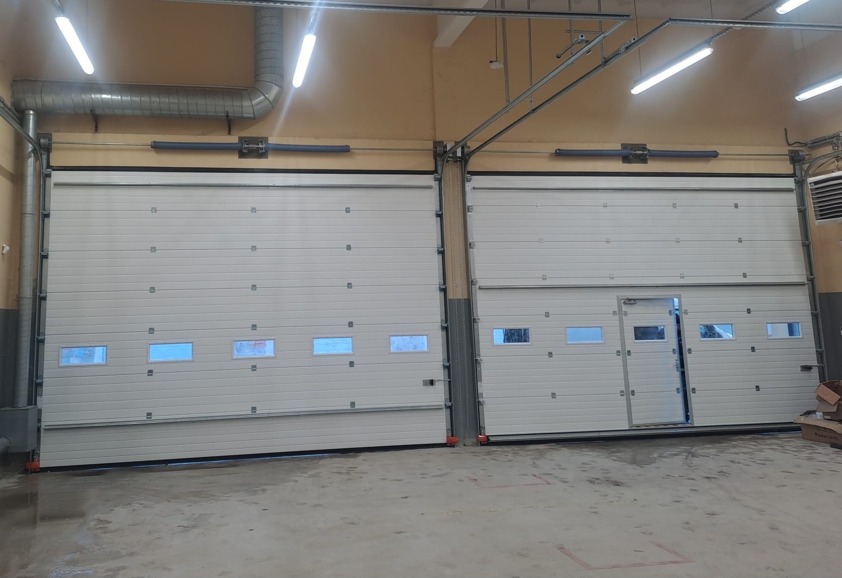 Dwie białe, segmentowe bramy garażowe z oknami, jedna z drzwiami wejściowymi, w hali z beżowymi ścianami i widoczną instalacją pod sufitem.