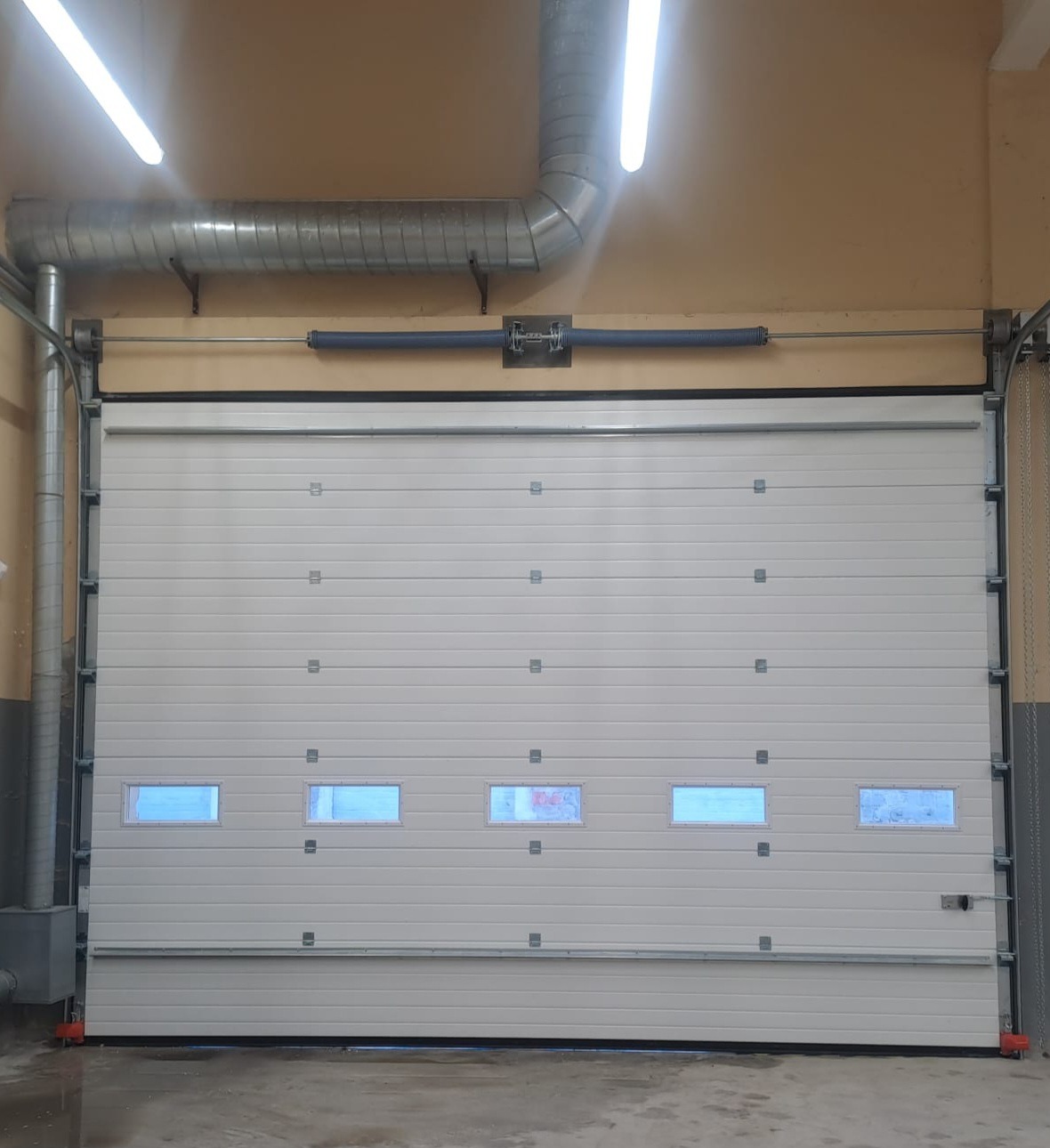 Zamknięta, segmentowa brama garażowa w kolorze białym z czterema małymi okienkami, widoczna wewnątrz budynku z instalacją wentylacyjną pod sufitem.