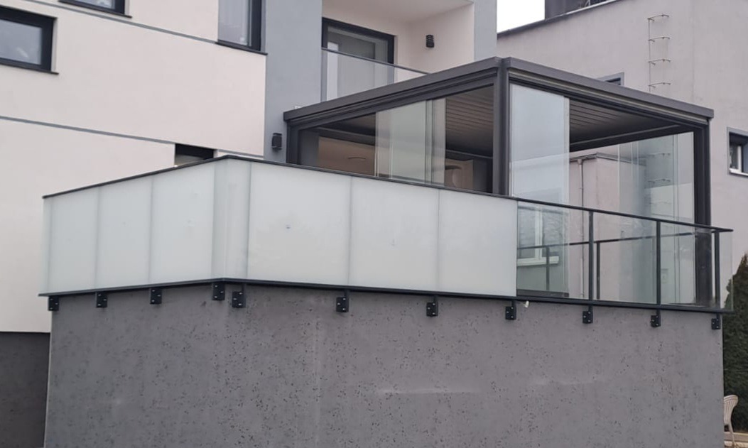 Nowoczesny balkon z przeszkloną zabudową i balustradą ze szkła hartowanego, czarne elementy konstrukcyjne, szara elewacja budynku.