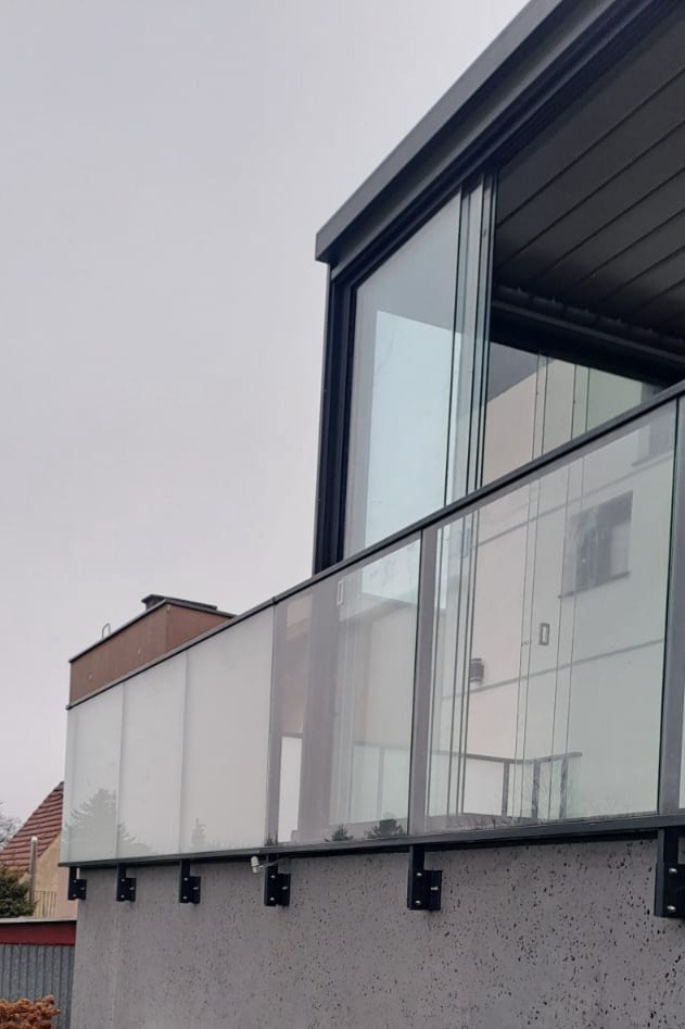 Szklana balustrada balkonowa z czarnymi mocowaniami, widok z dołu na tle szarego nieba i fragmentu budynku. Nowoczesny design.