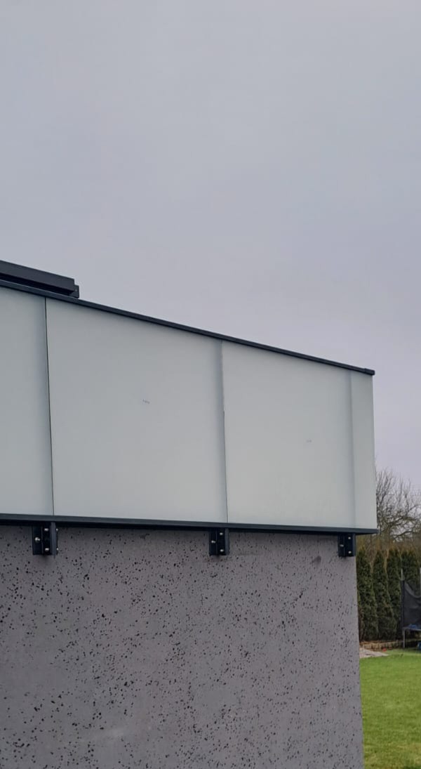 Nowoczesna balustrada szklana z czarnymi mocowaniami na szorstkim, betonowym murku. Widok z dołu, pochmurne niebo w tle.