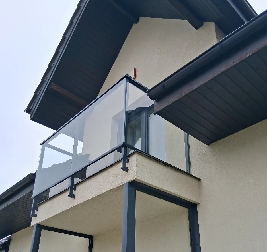 Szklana balustrada balkonowa z czarnymi elementami mocującymi, widok z dołu, na tle beżowej elewacji budynku z ciemną podbitką dachową.
