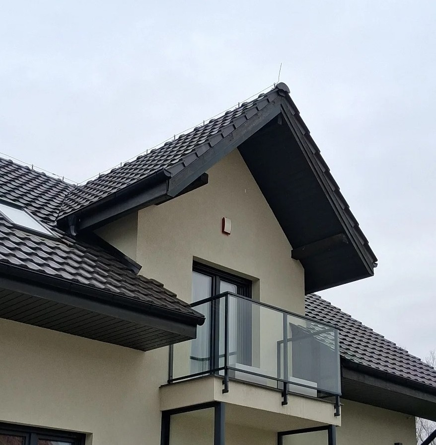 Nowoczesny balkon z czarną ramą i szklaną balustradą, widoczny z dołu, na tle beżowej elewacji domu z ciemną dachówką. Minimalistyczny design.