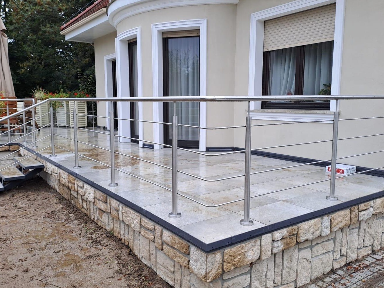 Błyszcząca, metalowa balustrada z poziomymi linkami na tarasie z kamienną podmurówką i jasnymi płytkami. Widok na elewację domu z oknami.