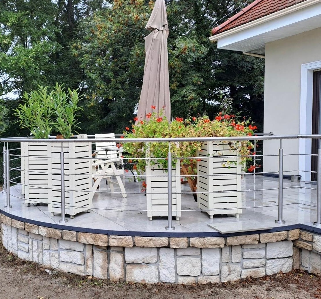 Balkon z niską, metalową balustradą, wykończony kamieniem. Na balkonie białe donice z kwiatami i krzesła. W tle zieleń i fragment budynku z beżową elewacją.