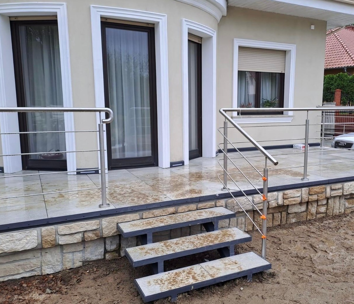 Nowoczesna balustrada ze stali nierdzewnej przy wejściu do domu z kamienną elewacją i schodami. Widoczne okna z białymi ramami i brązowymi roletami.