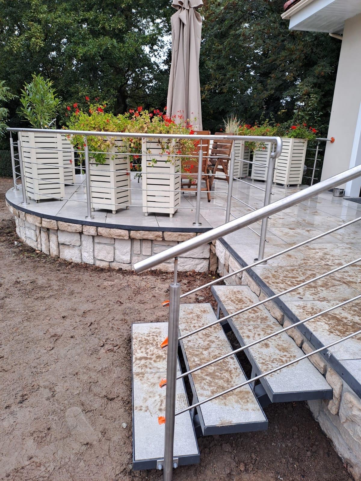 Taras z balustradą ze stali nierdzewnej i schodami z kamiennymi stopniami. Na tarasie donice z kwiatami i parasol. Widok na ogród.