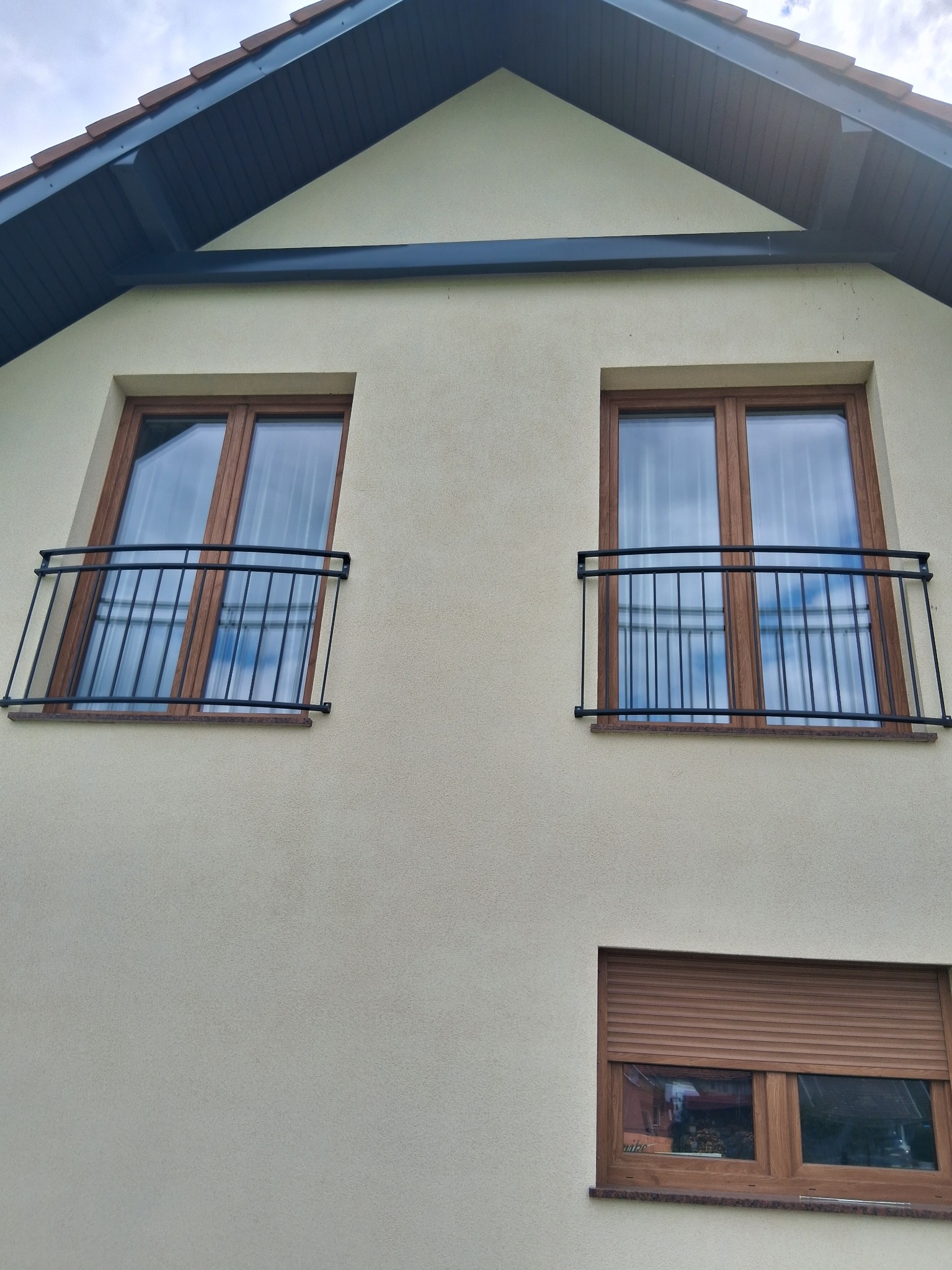 Elewacja domu z dwoma oknami na piętrze, każde z czarną, metalową balustradą balkonową. Na dole okno z brązowymi roletami zewnętrznymi.