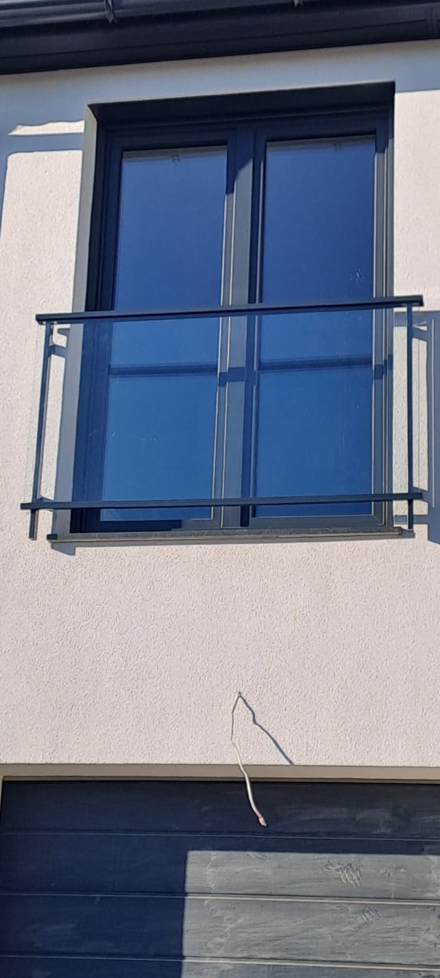 Szklana balustrada balkonowa z czarnymi, metalowymi elementami mocującymi, zamontowana na elewacji budynku nad garażem. Widok z dołu.