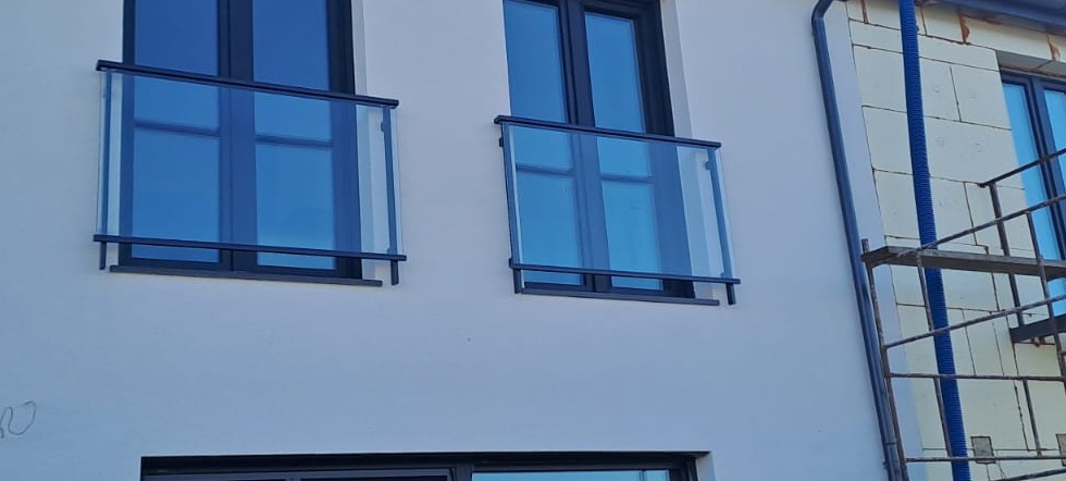Elewacja budynku z dwoma szklanymi balustradami balkonowymi z czarnymi ramami, widoczne rusztowanie z boku. Minimalistyczny design.
