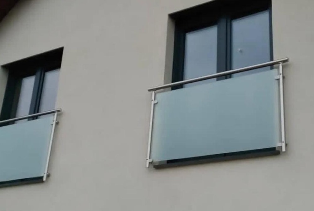 Dwa balkony z matowym szkłem i metalowymi poręczami, zamontowane na jasnej elewacji budynku. Widok z dołu, symetryczny układ okien i balkonów.