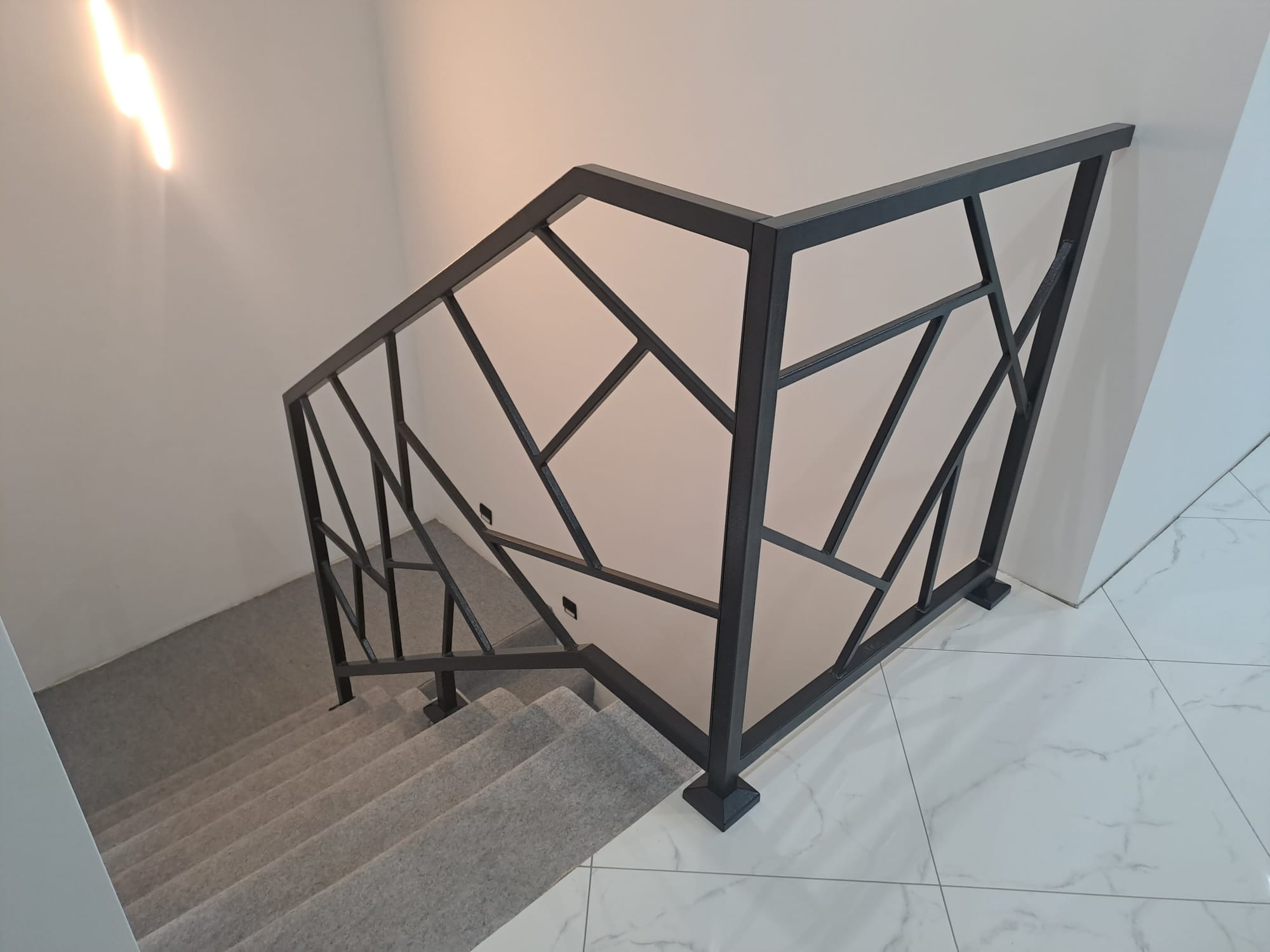 Nowoczesna, czarna poręcz schodowa z geometrycznym wzorem, zamontowana w jasnym wnętrzu. Widok z góry na schody pokryte wykładziną i marmurową podłogę.