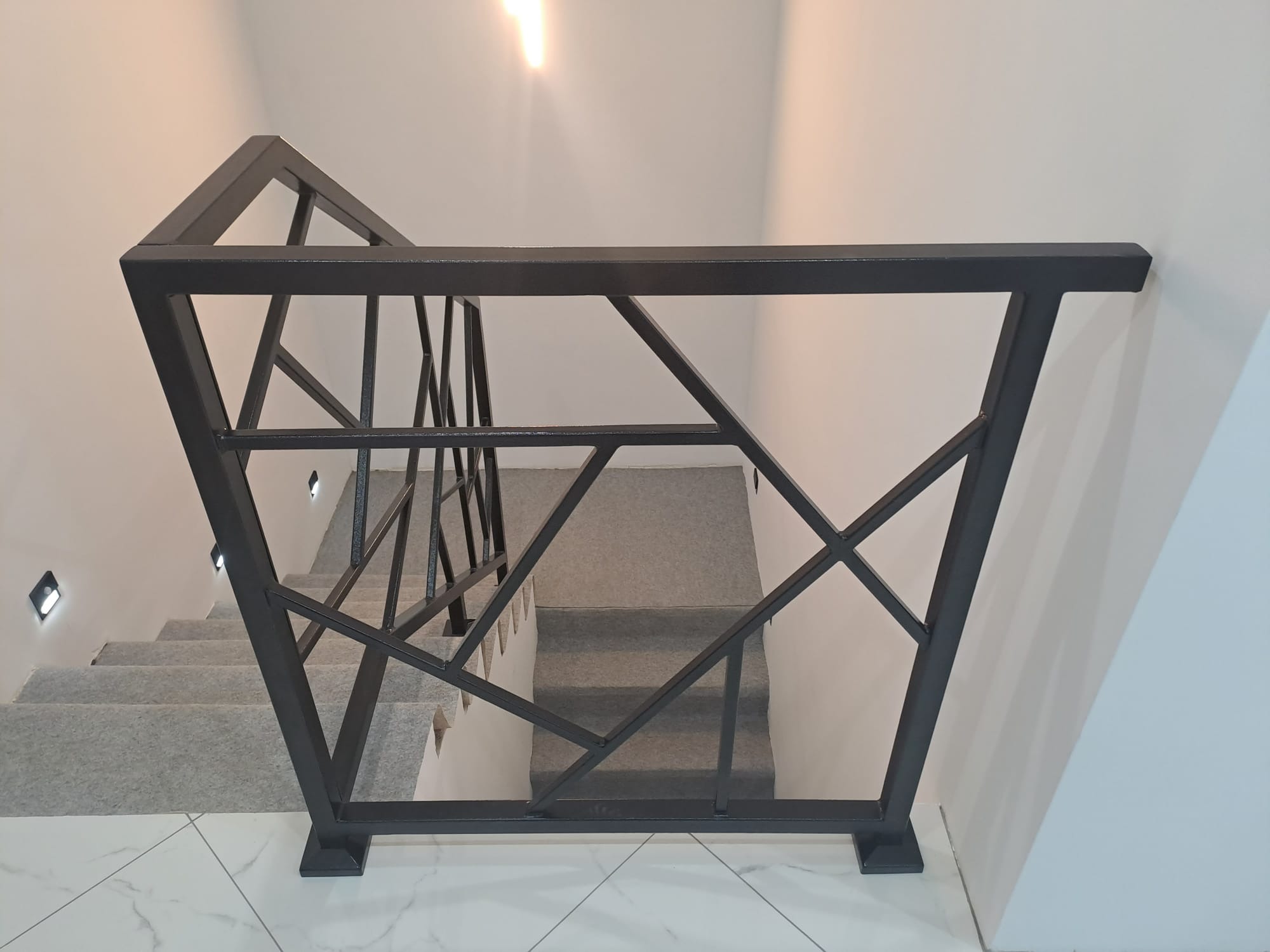 Czarna, metalowa balustrada schodowa o geometrycznym wzorze, widziana z góry. Schody wyłożone szarą wykładziną, białe ściany i podłoga z płytek imitujących marmur.
