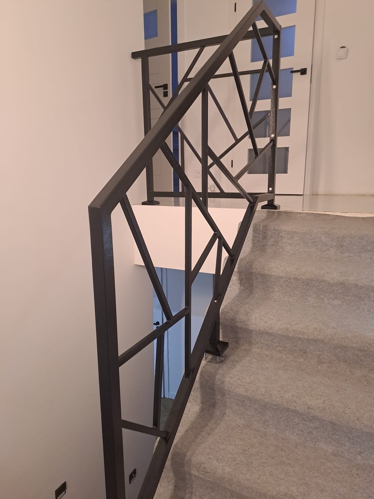 Nowoczesna poręcz schodowa z geometrycznym wzorem, wykonana z ciemnego metalu. Widok z góry na schody pokryte wykładziną. W tle białe ściany i drzwi.