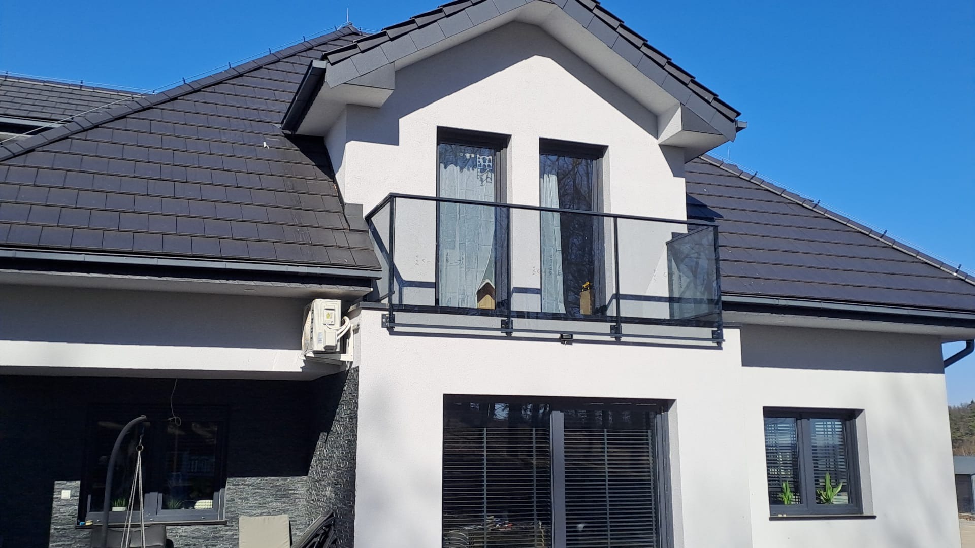 Nowoczesny balkon z czarną, szklaną balustradą na tle białej fasady domu z ciemną dachówką i błękitnym niebem. Widok z dołu, podkreślający minimalistyczny design.