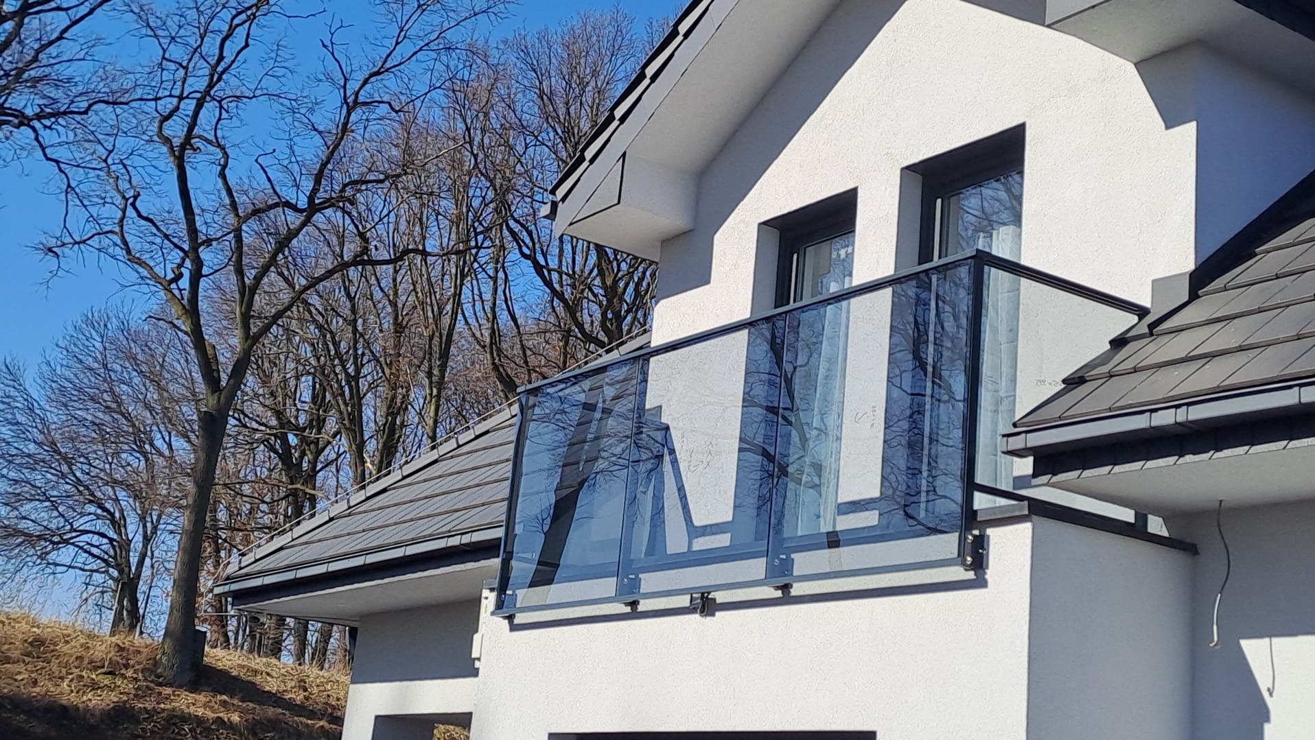 Szklana balustrada balkonowa z czarnymi profilami, zamontowana na białym budynku z ciemną dachówką. W tle widoczne drzewa i błękitne niebo.