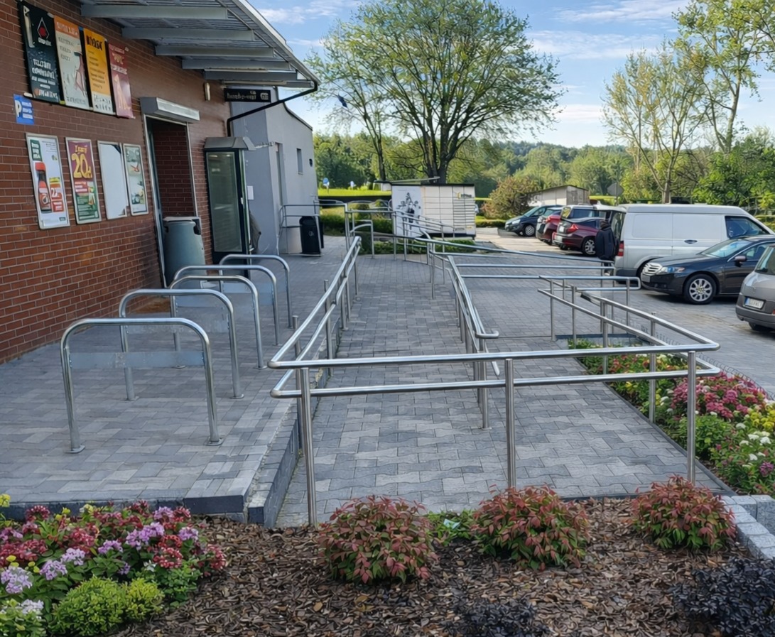 Zewnętrzny podjazd dla niepełnosprawnych z metalowymi poręczami, prowadzący do budynku z cegły. Widoczne zaparkowane samochody i zieleń w tle.