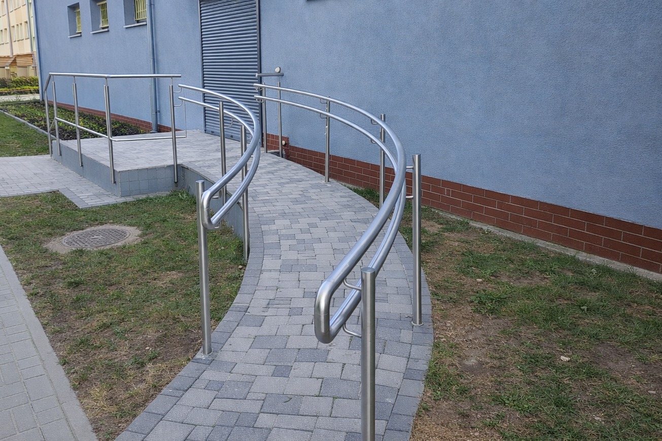Zakrzywiona ścieżka z szarej kostki brukowej, otoczona trawnikiem, z zamontowanymi po obu stronach metalowymi poręczami. Budynek w tle.