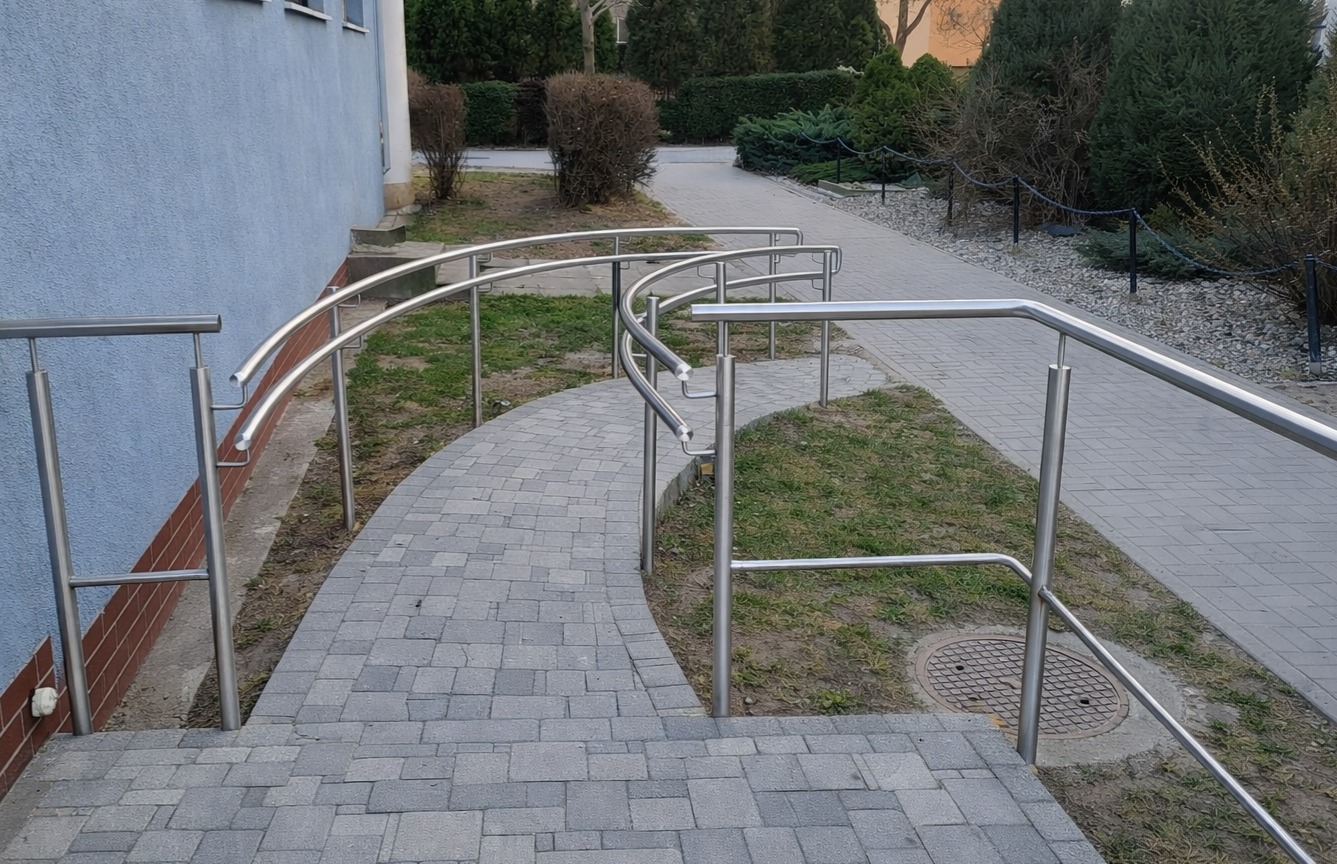 Zakrzywione poręcze ze stali nierdzewnej wzdłuż ścieżki z kostki brukowej, prowadzącej do budynku. Widoczne krzewy i trawa. Ujęcie z perspektywy schodów.