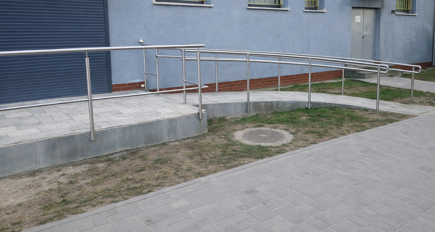 Zewnętrzna rampa z poręczami ze stali nierdzewnej, zapewniająca dostęp do budynku. Widoczne szare płytki chodnikowe i fragment elewacji budynku w odcieniach szarości i błękitu.