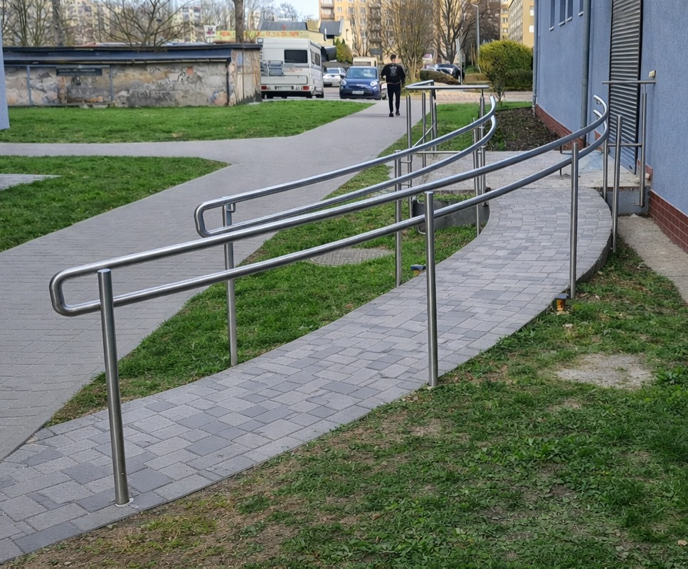 Zewnętrzne, metalowe poręcze przy chodniku z kostki brukowej, ułatwiające dostęp do budynku. W tle osoba idąca chodnikiem i zaparkowane pojazdy.