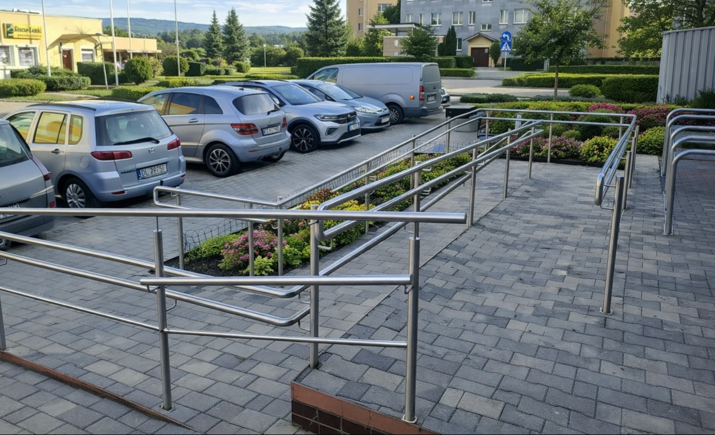 Zewnętrzne, metalowe poręcze przy chodniku z kostki brukowej, z rampą dla wózków. W tle zaparkowane samochody i budynek.