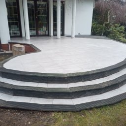 Mariusz Markowski - Wyłożony jasnymi płytkami taras z trzema stopniami wykończonymi ciemnym drewnem, widok z dołu na dom z dużymi oknami.