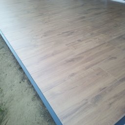 Mariusz Markowski - Ułożona podłoga z płytek imitujących drewno na zewnątrz budynku, widoczne szare obrzeże i fragment elewacji z oknami przesuwnymi.