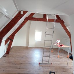 Mariusz Markowski - Poddasze po malowaniu z drewnianymi belkami, aluminiowa drabina oparta o ścianę, stolik z farbami, surowe oświetlenie żarówką
